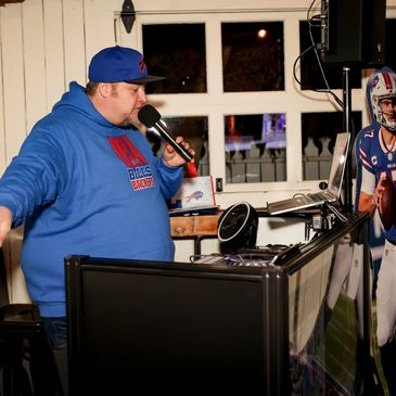 Man in a blue hoodie and cap speaking into a microphone at a DJ setup.